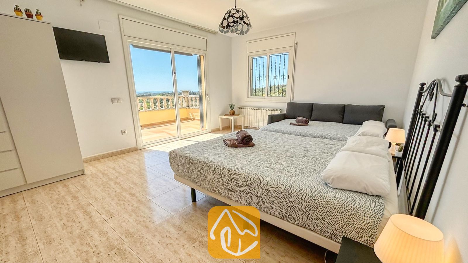 Vakantievilla's Costa Brava Spanje - Villa Holiday - Slaapkamer met twee bedden, tegelvloeren, een wandtelevisie en glazen schuifdeuren die uitkomen op een zonnig balkon.