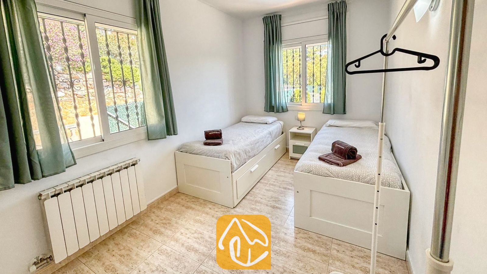 Vakantievilla's Costa Brava Spanje - Villa Holiday - Slaapkamer met twee witte eenpersoonsbedden, opgerolde bruine handdoeken, groene gordijnen en tegelvloeren.
