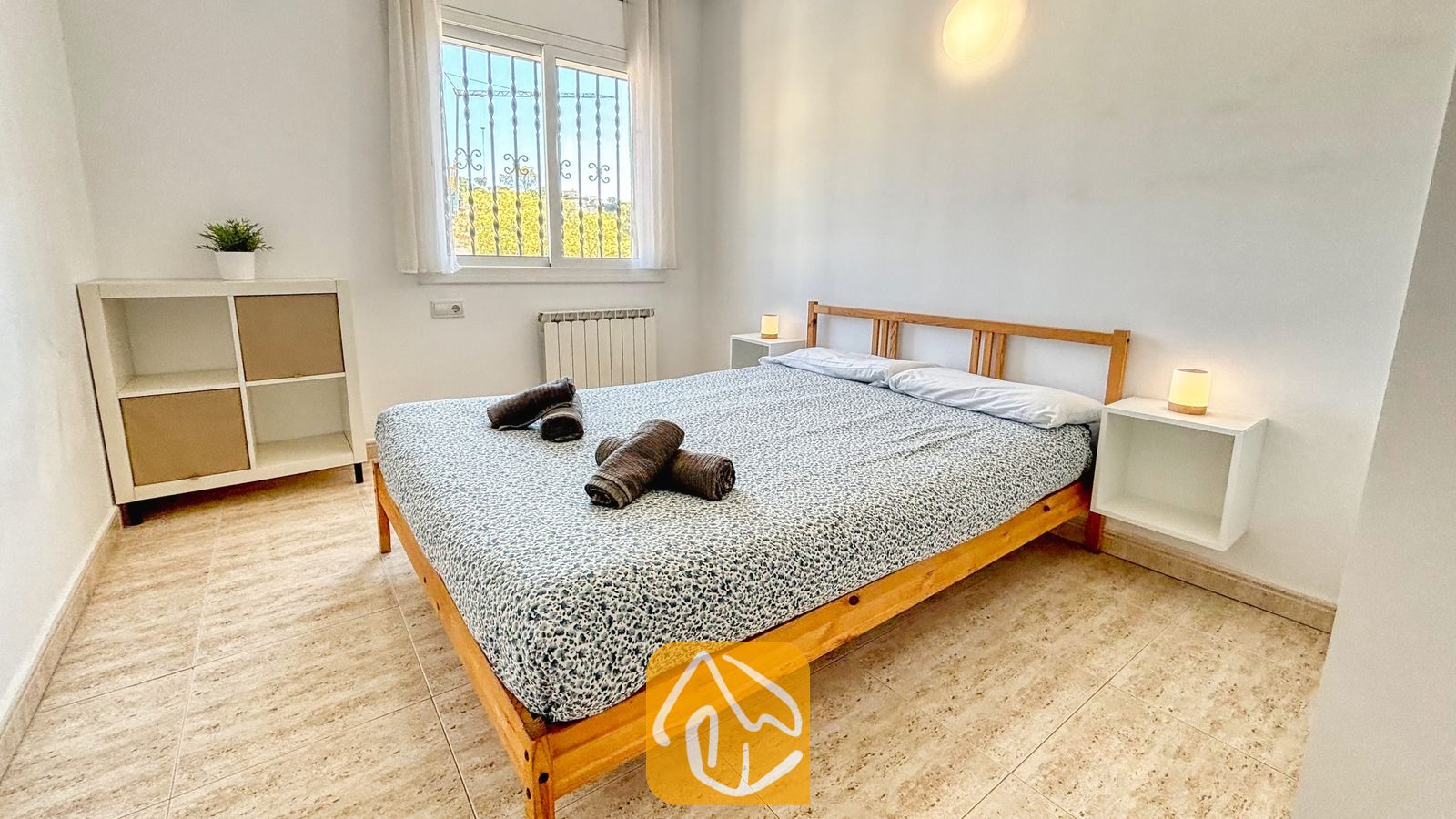 Vakantievilla's Costa Brava Spanje - Villa Holiday - Slaapkamer met een houten bedframe, patroon in grijs beddengoed en opgerolde donkere handdoeken erop, geflankeerd door twee kleine witte nachtkastjes met lampen.
