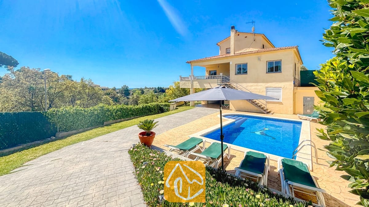 Vakantievilla's Costa Brava Spanje - Villa Holiday - Buitenaanzicht van Villa Holiday met het gele huis van twee verdiepingen, het zwembad met dolfijnmozaïek en groene ligstoelen onder een grijze parasol.
