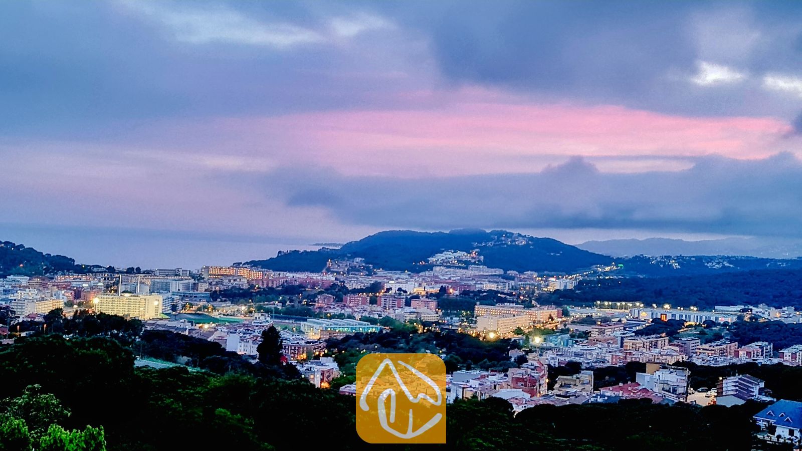 Vakantievilla's Costa Brava Spanje - Villa Maribel - Verheven uitzicht vanuit Villa Maribel over een stad en heuvels onder een bewolkte schemeringshemel met roze en blauwe tinten.
