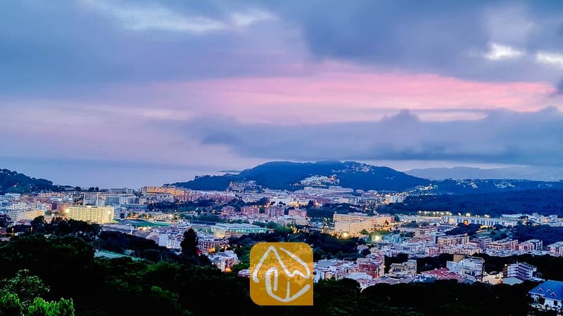 Vakantievilla's Costa Brava Spanje - Villa Maribel - Verheven uitzicht vanuit Villa Maribel over een stad en heuvels onder een bewolkte schemeringshemel met roze en blauwe tinten.