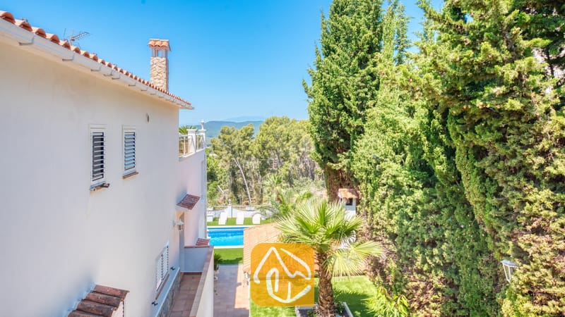Vakantievilla's Costa Brava Spanje - Villa Maribel - Buitenaanzicht van Villa Maribel met een wit gepleisterde muur voorzien van luiken, terracotta dakpannen en dichte groene cipressen die een fraai omlijst uitzicht bieden op het verre landschap en het zwembad.