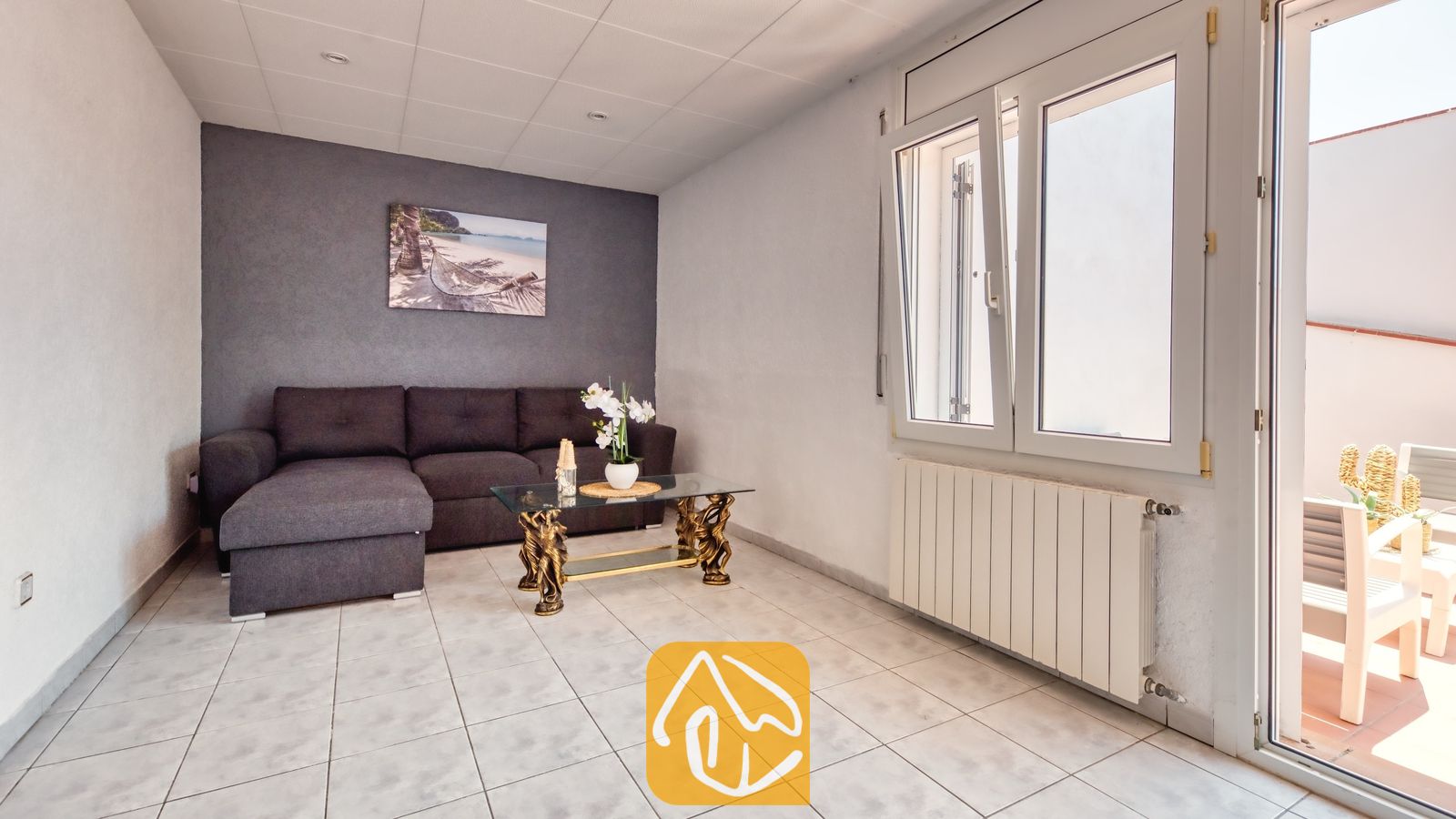 Vakantievilla's Costa Brava Spanje - Villa Maribel - Interieur van de woonkamer in Villa Maribel met een donkergrijze hoekbank, een glazen salontafel met sierlijke gouden poten en een tegelvloer.