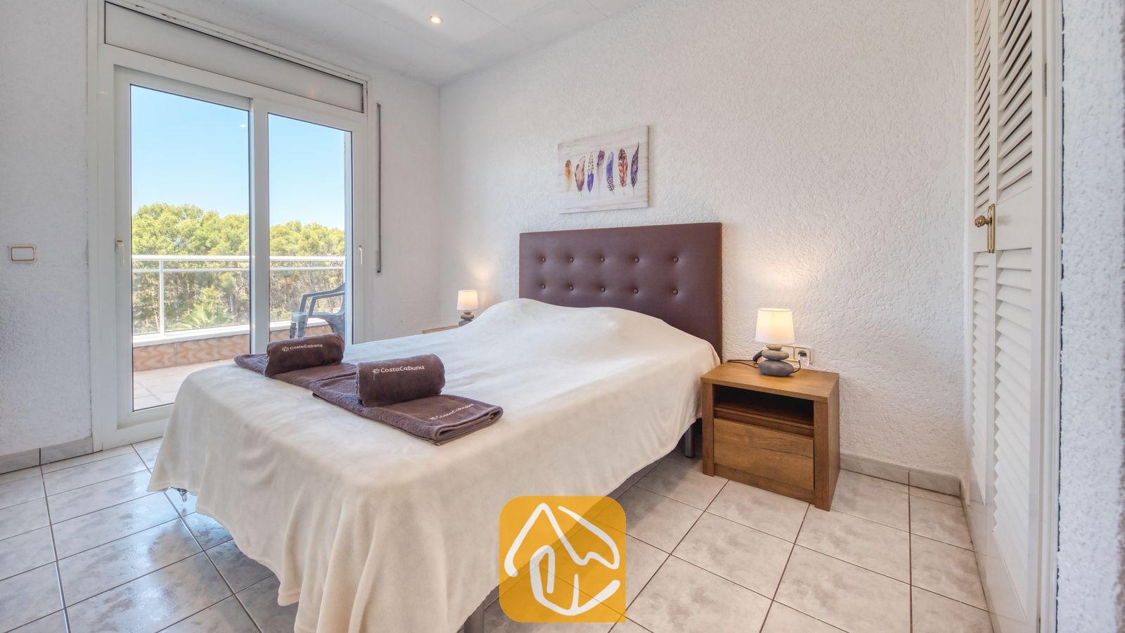 Vakantievilla's Costa Brava Spanje - Villa Maribel - Slaapkamer met een bruin, getufte hoofdbord, opgerolde handdoeken op het bed en een glazen schuifdeur die uitkomt op een balkon met uitzicht op de bomen.