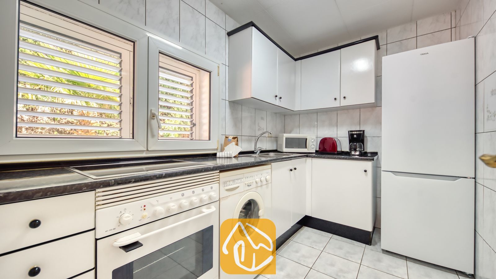Vakantievilla's Costa Brava Spanje - Villa Maribel - Keuken met witte kasten, een ingebouwde oven, wasmachine en twee ramen met rolluiken aan de buitenzijde.