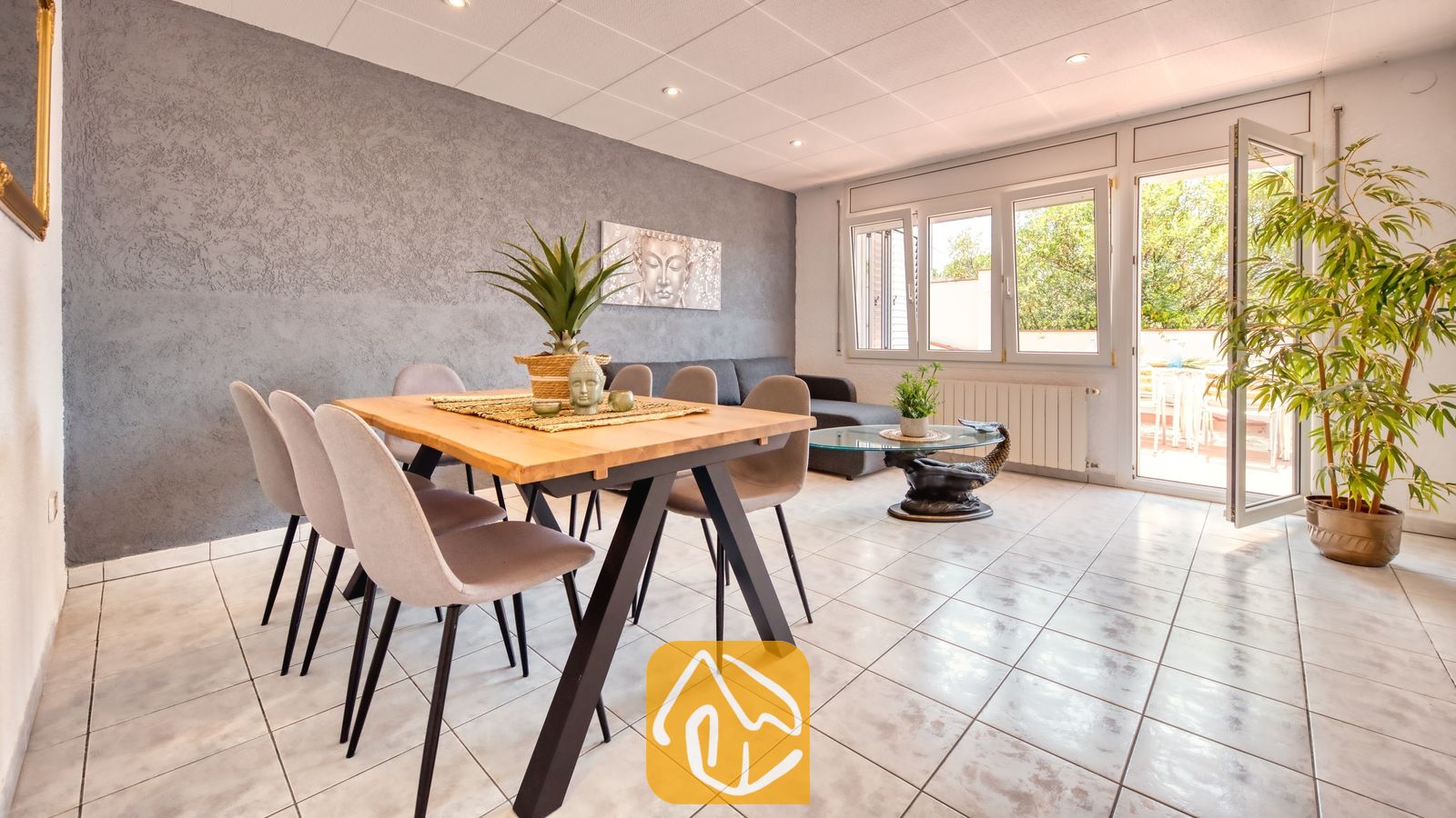 Vakantievilla's Costa Brava Spanje - Villa Maribel - Eethoek met een houten tafel, gestoffeerde stoelen en grijs getextureerde muren in Villa Maribel, voorzien van glazen deuren die uitkomen op een terras.