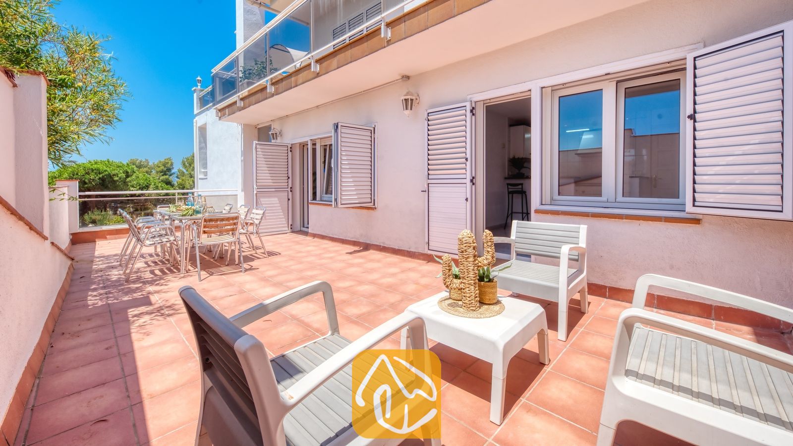 Vakantievilla's Costa Brava Spanje - Villa Maribel - Zonnig buitenterras bij Villa Maribel met betegelde vloeren, witte plastic stoelen, een kleine salontafel en open ramen met luiken.