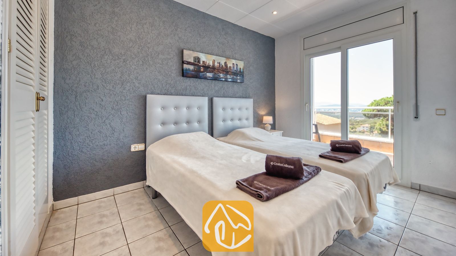 Vakantievilla's Costa Brava Spanje - Villa Maribel - Slaapkamer met twee eenpersoonsbedden tegen een getextureerde grijze wand, tegelvloer en schuifpui die toegang biedt tot een balkon.