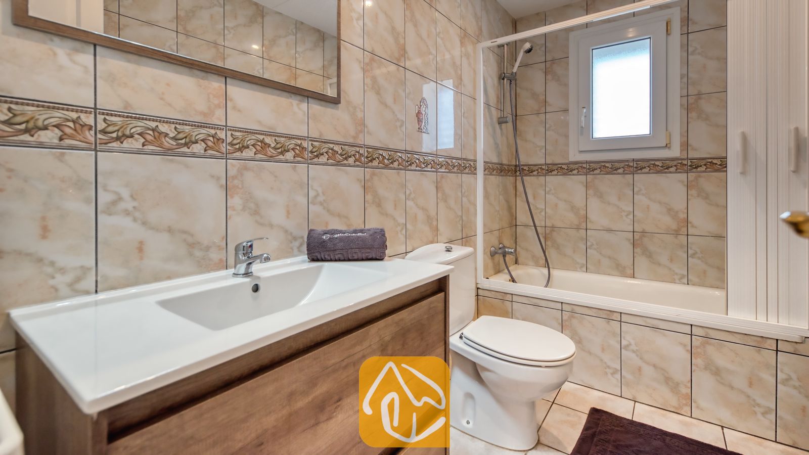 Vakantievilla's Costa Brava Spanje - Villa Maribel - Badkamer voorzien van een witte keramische wastafel met houten meubel, een toilet en een ligbad met doucheschep, omringd door beige marmerlook tegels.