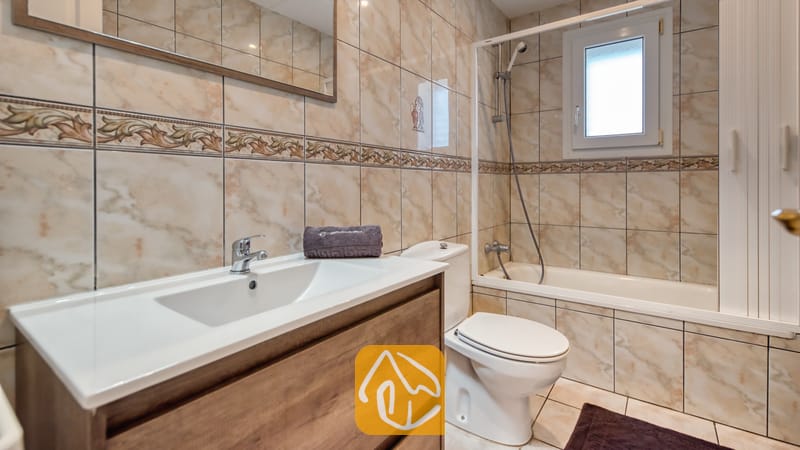 Vakantievilla's Costa Brava Spanje - Villa Maribel - Badkamer voorzien van een witte keramische wastafel met houten meubel, een toilet en een ligbad met doucheschep, omringd door beige marmerlook tegels.
