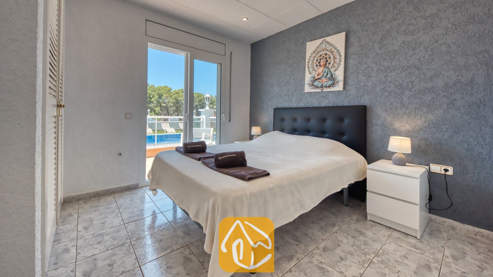Vakantievilla's Costa Brava Spanje - Villa Maribel - Slaapkamer met een zwart gestoffeerd hoofdbord, witte nachtkastjes met lampen en glazen deuren die uitkomen op een terras met uitzicht op het zwembad.