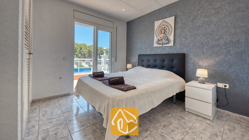 Vakantievilla's Costa Brava Spanje - Villa Maribel - Slaapkamer met een zwart gestoffeerd hoofdbord, witte nachtkastjes met lampen en glazen deuren die uitkomen op een terras met uitzicht op het zwembad.