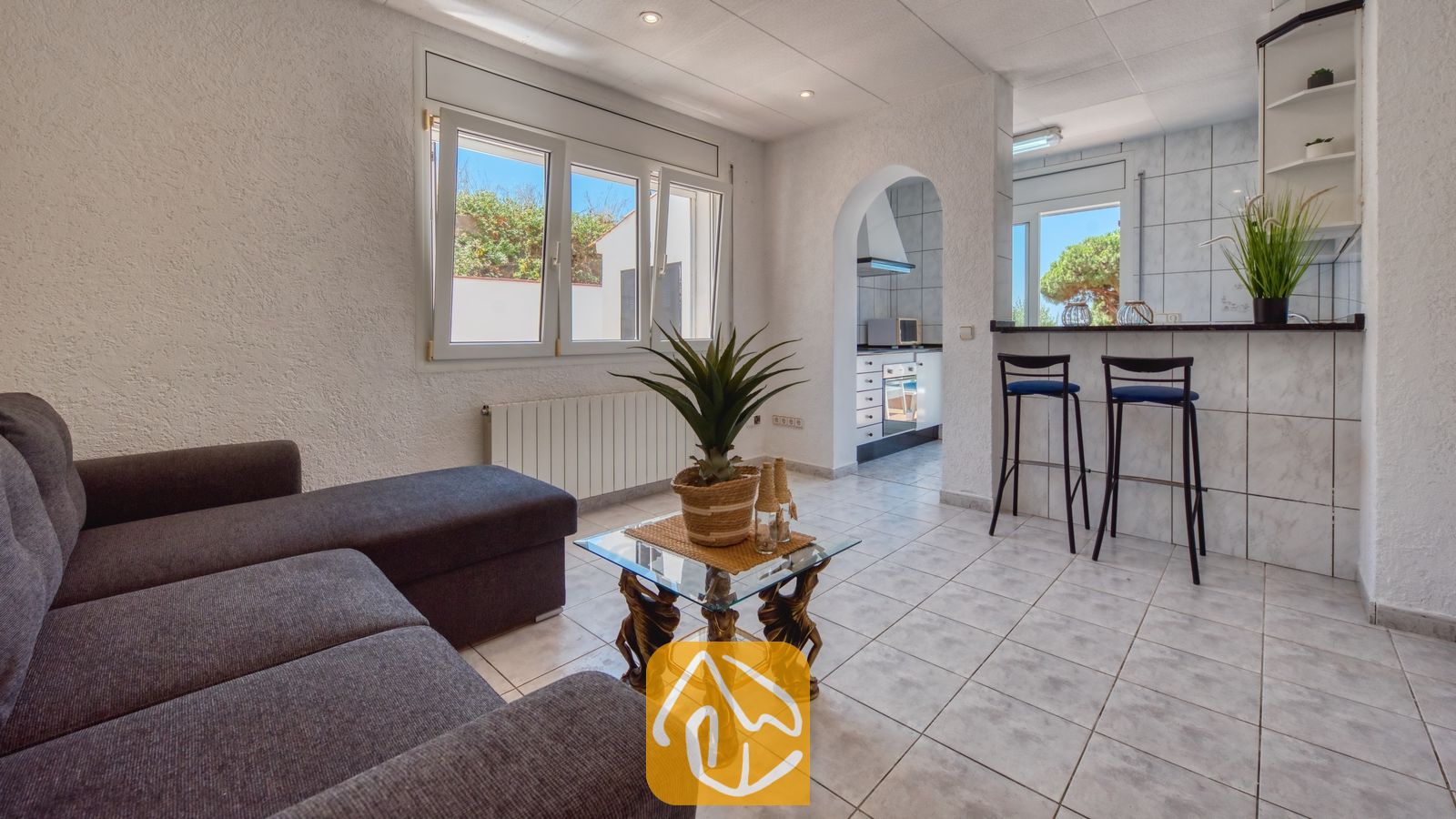 Vakantievilla's Costa Brava Spanje - Villa Maribel - Woonkamer met een grijze hoekbank, een salontafel met glazen blad en een open doorgang naar de keuken met barstoelen in Villa Maribel.