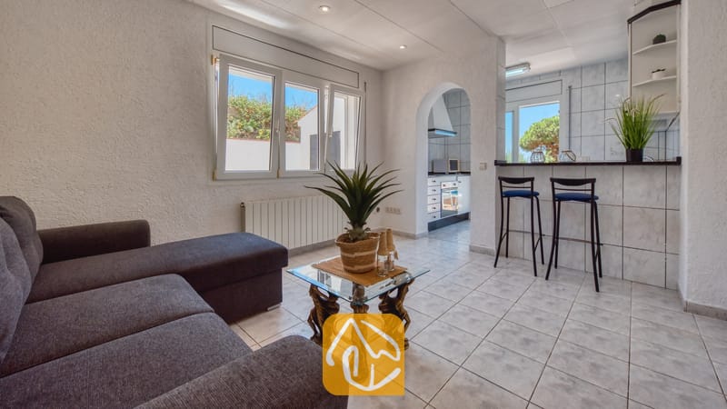 Vakantievilla's Costa Brava Spanje - Villa Maribel - Woonkamer met een grijze hoekbank, een salontafel met glazen blad en een open doorgang naar de keuken met barstoelen in Villa Maribel.