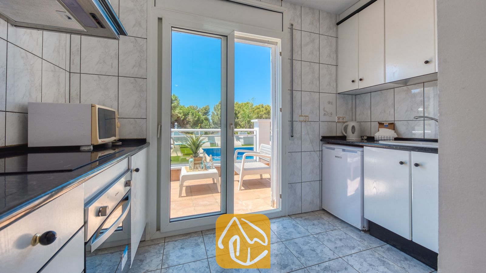 Vakantievilla's Costa Brava Spanje - Villa Maribel - Interieur van de keuken met witte kasten, betegelde muren en glazen schuifdeuren die uitkomen op een buitenterras met uitzicht op het zwembad bij Villa Maribel.