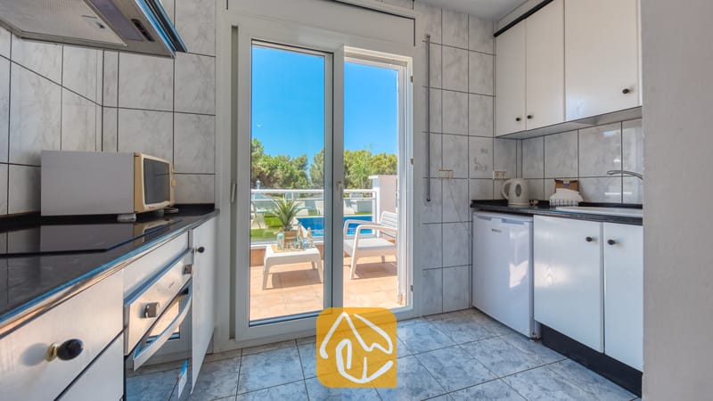 Vakantievilla's Costa Brava Spanje - Villa Maribel - Interieur van de keuken met witte kasten, betegelde muren en glazen schuifdeuren die uitkomen op een buitenterras met uitzicht op het zwembad bij Villa Maribel.