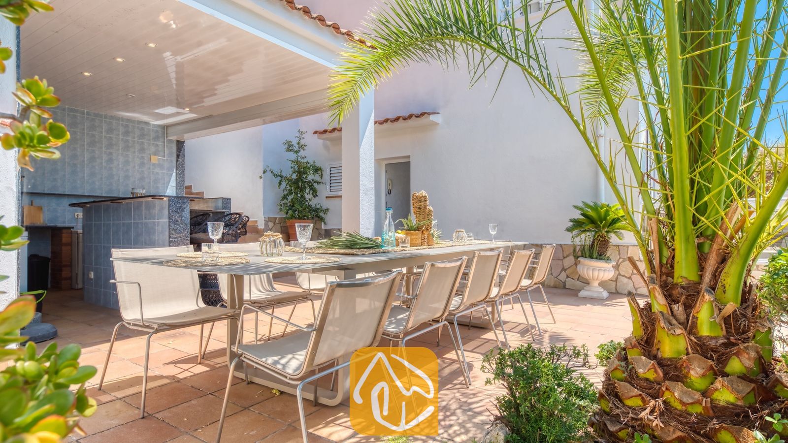 Vakantievilla's Costa Brava Spanje - Villa Maribel - Overdekt buitenterras om te dineren bij Villa Maribel, voorzien van een lange eettafel met stoelen, een betegelde bar en een groene palmboom op de voorgrond.