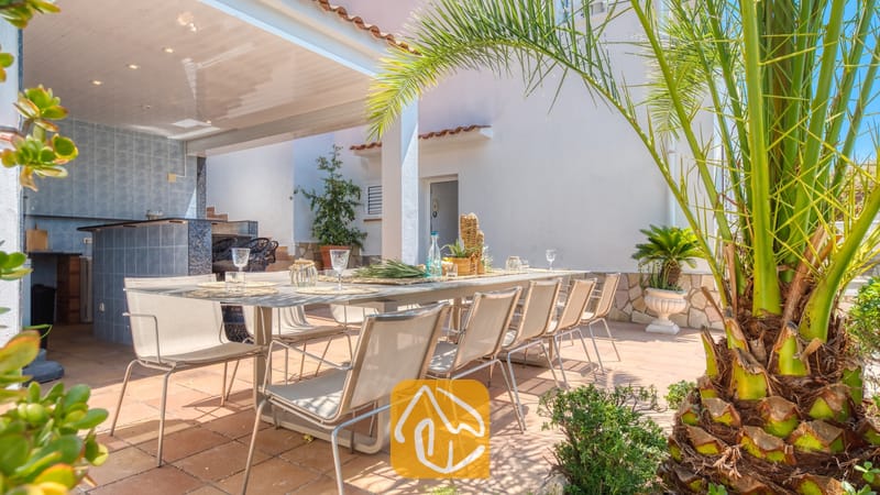 Vakantievilla's Costa Brava Spanje - Villa Maribel - Overdekt buitenterras om te dineren bij Villa Maribel, voorzien van een lange eettafel met stoelen, een betegelde bar en een groene palmboom op de voorgrond.