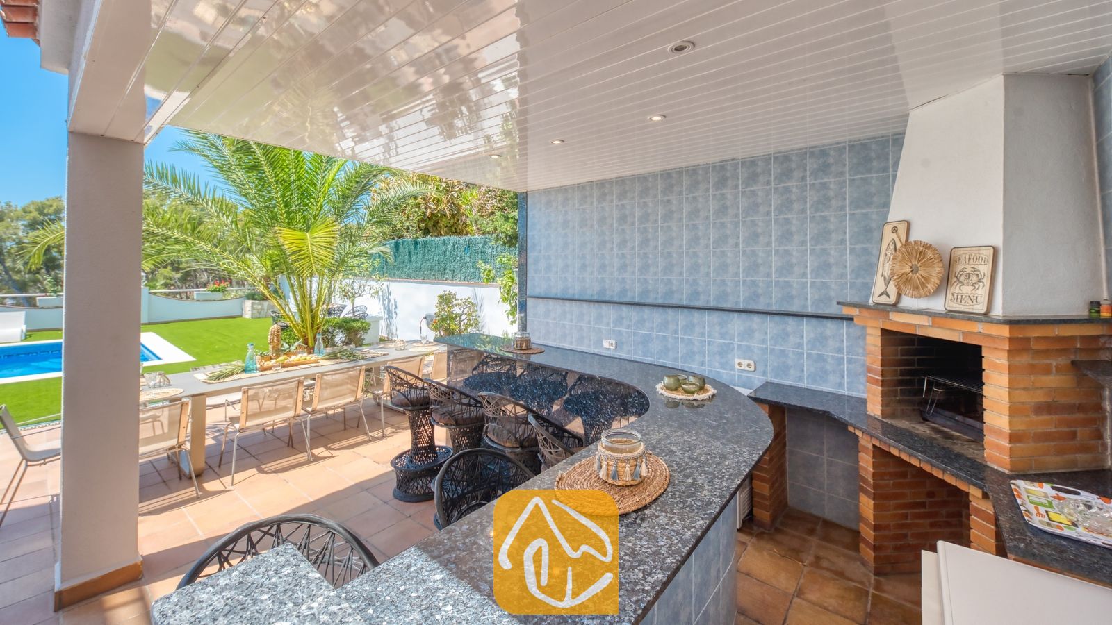 Vakantievilla's Costa Brava Spanje - Villa Maribel - Overdekte buitenbar en bakstenen barbecuehoek bij Villa Maribel, voorzien van granieten aanrechtbladen, blauw betegelde muren en rieten barstoelen met uitzicht op het zwembadterras.