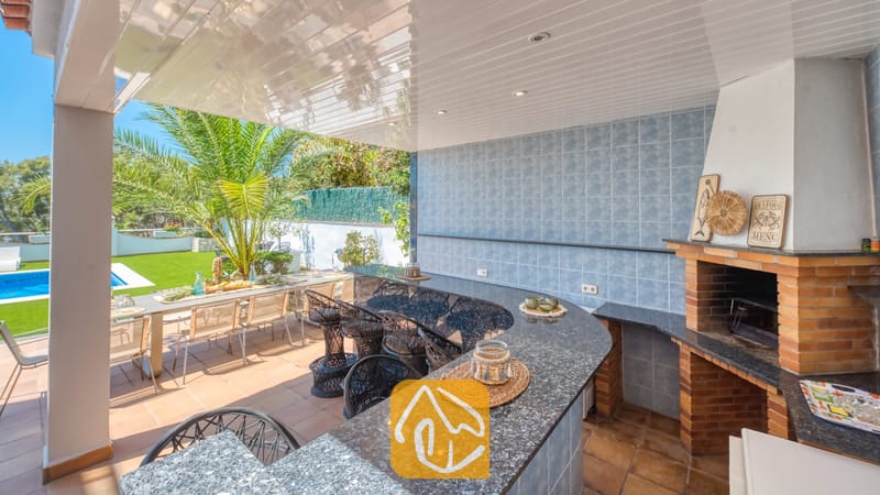 Vakantievilla's Costa Brava Spanje - Villa Maribel - Overdekte buitenbar en bakstenen barbecuehoek bij Villa Maribel, voorzien van granieten aanrechtbladen, blauw betegelde muren en rieten barstoelen met uitzicht op het zwembadterras.