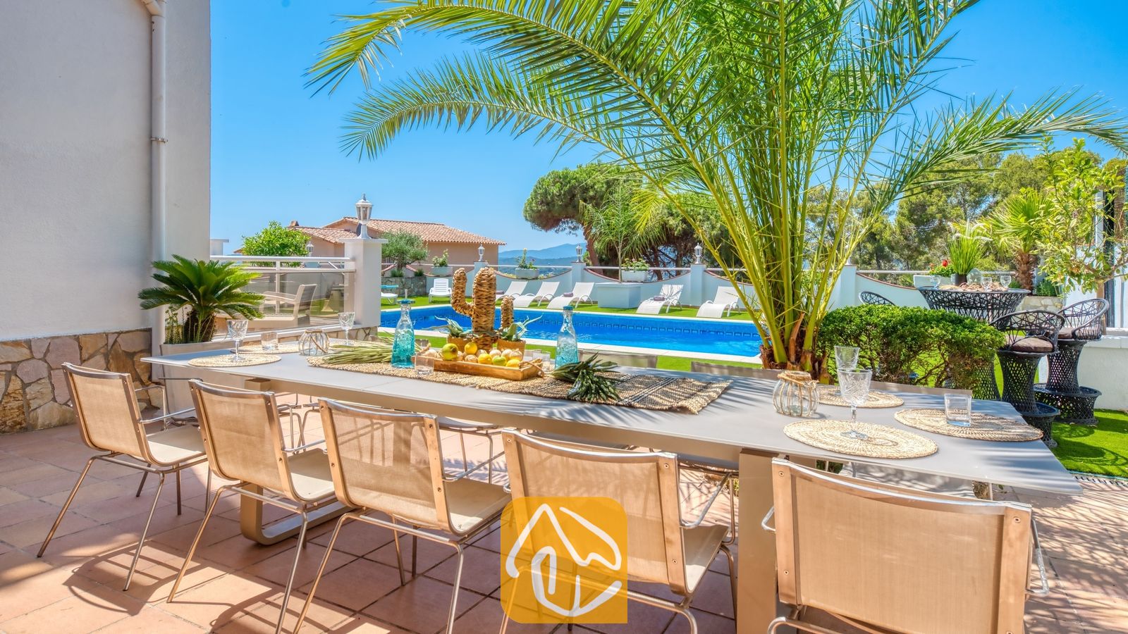 Vakantievilla's Costa Brava Spanje - Villa Maribel - Villa Maribel terras met een lange eettafel voor buiten, beige stoelen van gaas en uitzicht op het zwembad en de palmboom.