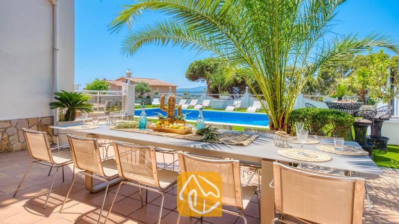 Vakantievilla's Costa Brava Spanje - Villa Maribel - Villa Maribel terras met een lange eettafel voor buiten, beige stoelen van gaas en uitzicht op het zwembad en de palmboom.