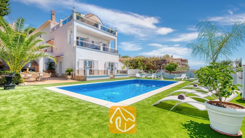 Vakantievilla's Costa Brava Spanje - Villa Maribel - Privézwembad en witte ligbedden op een groen gazon bij Villa Maribel in Spanje, met een wit villa met meerdere verdiepingen, balkons en palmbomen.