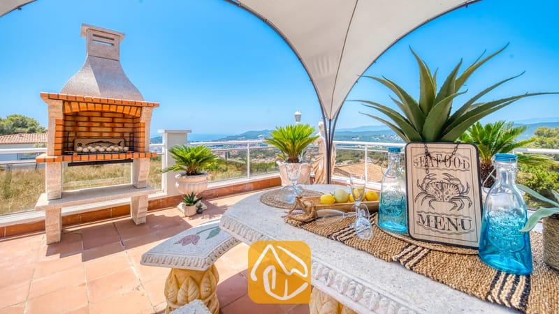 Vakantievilla's Costa Brava Spanje - Villa Maribel - Overdekt terras bij Villa Maribel met een stenen eethoek, een stenen barbecue en een ver uitzicht op zee onder een helderblauwe hemel.
