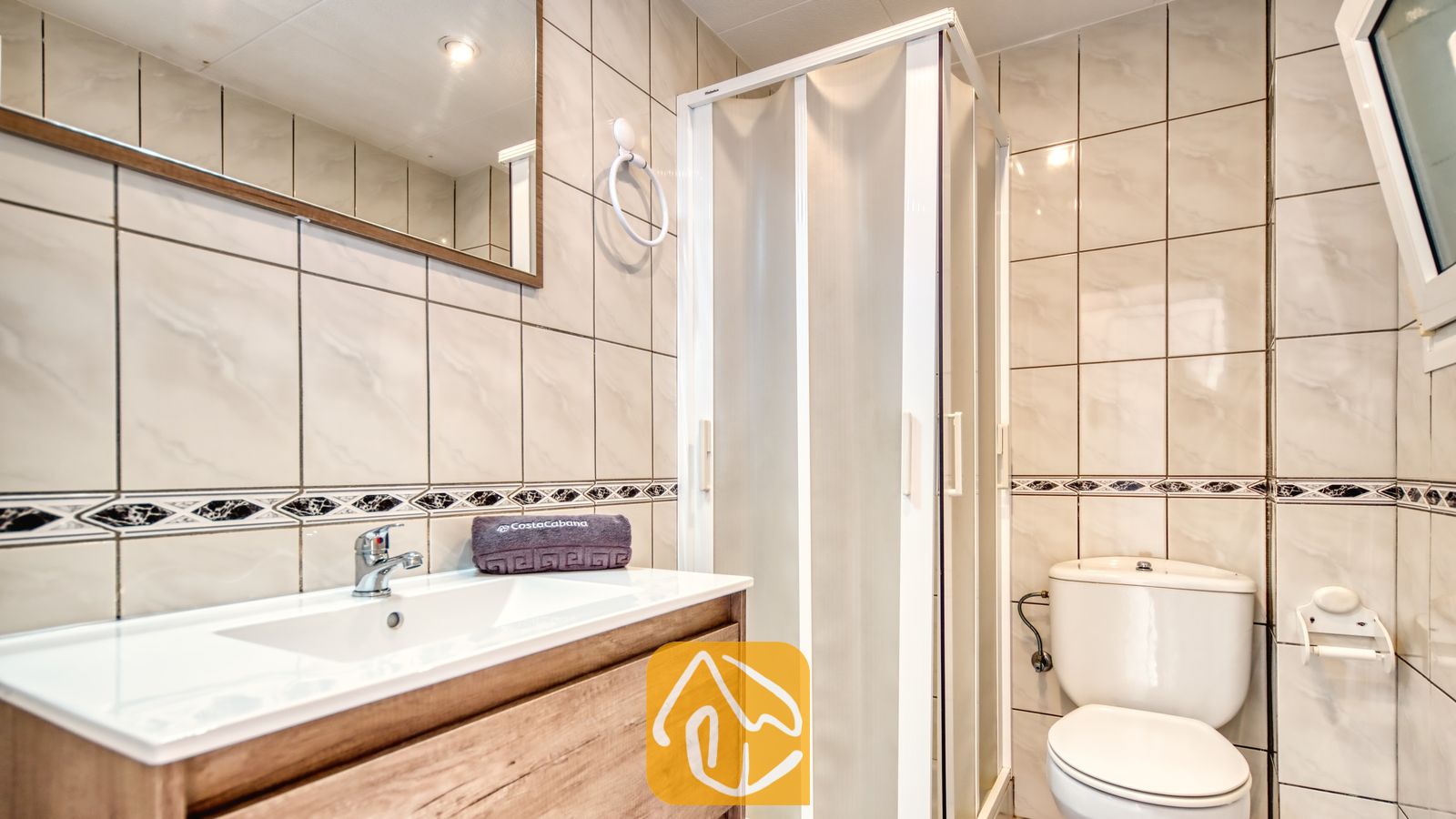 Vakantievilla's Costa Brava Spanje - Villa Maribel - Badkamer met beige wandtegels, een houten badkamermeubel met een witte wastafel, een hoekdouche en een toilet.