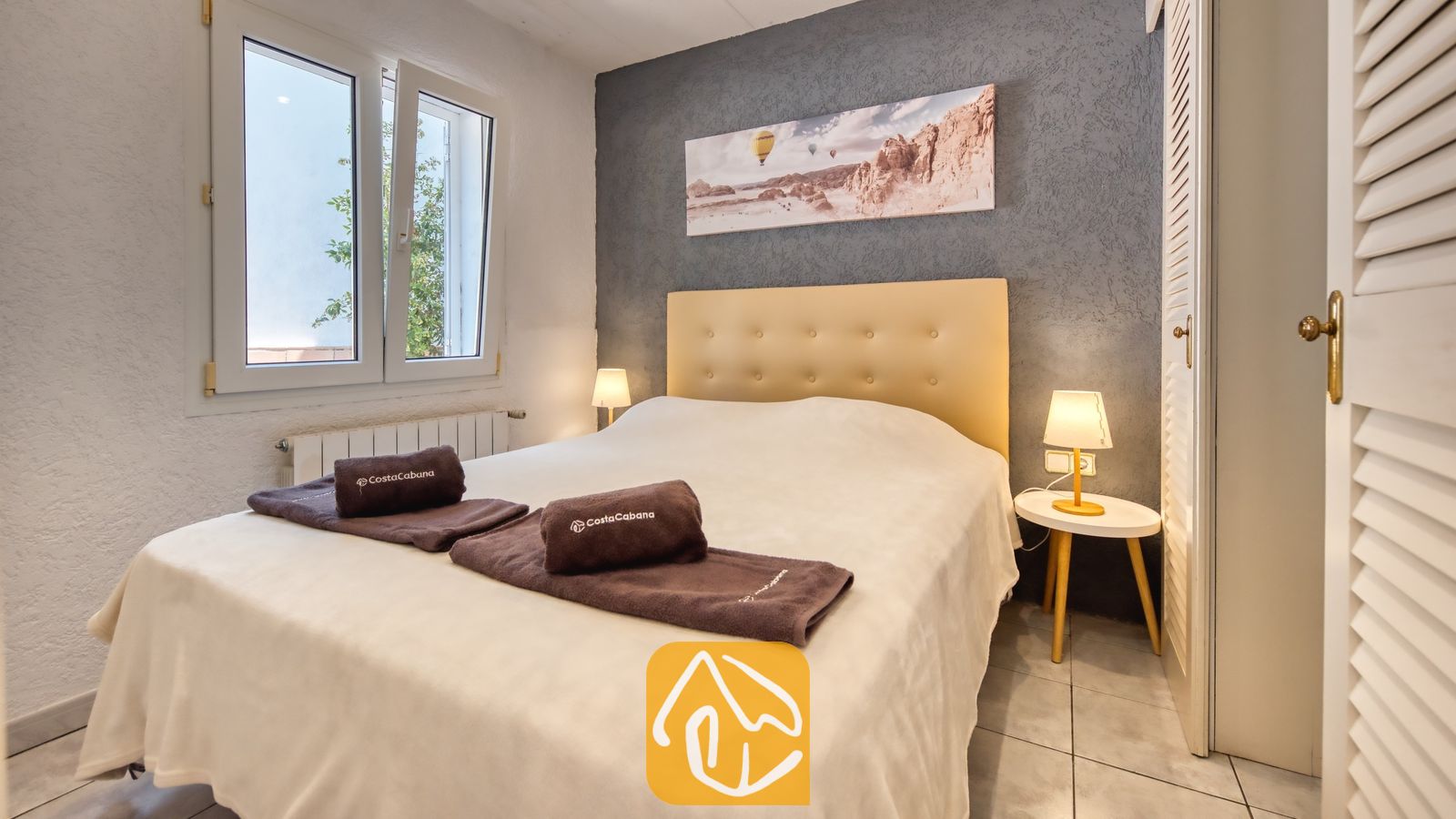 Vakantievilla's Costa Brava Spanje - Villa Maribel - Slaapkamer met een tweepersoonsbed, witt linnen, bruine opgerolde handdoeken en een getextureerde donkergrijze accentmuur onder een canvas landschapsschilderij.