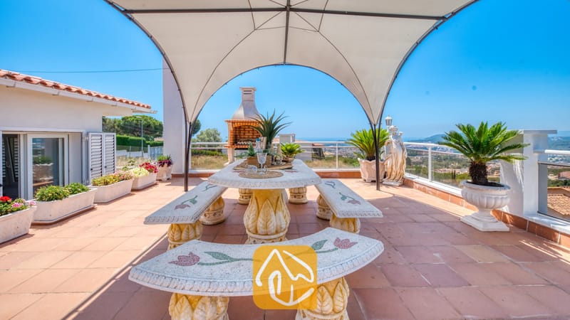 Vakantievilla's Costa Brava Spanje - Villa Maribel - Overdekt terras bij Villa Maribel met een stenen eettafel en bijpassende gebogen banken, potplanten en een stenen barbecue in de open lucht met uitzicht op een verafgelegen landschap.