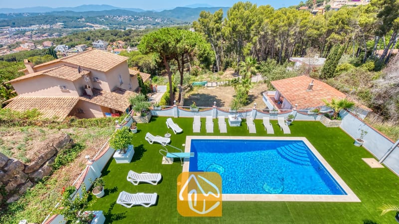Vakantievilla's Costa Brava Spanje - Villa Maribel - Luchtfoto van Villa Maribel met een rechthoekig buitenzwembad, omringd door witte ligstoelen op een groen gazon, met terracotta dakpannen en heuvels in de verte.