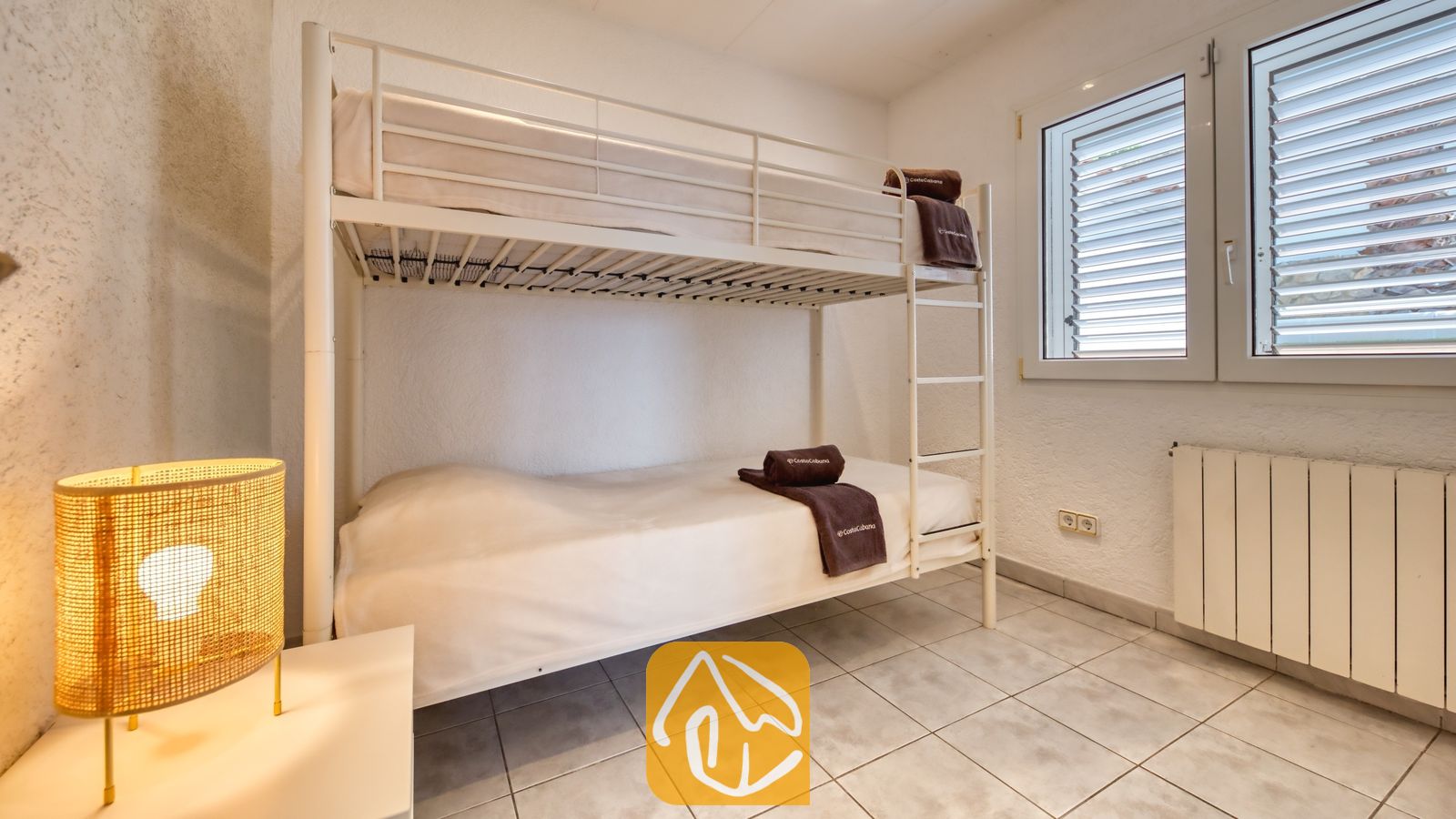 Vakantievilla's Costa Brava Spanje - Villa Maribel - Slaapkamer met een wit metalen stapelbed, een tegelvloer, een geweven lamp op een nachtkastje en twee ramen met witte luiken.