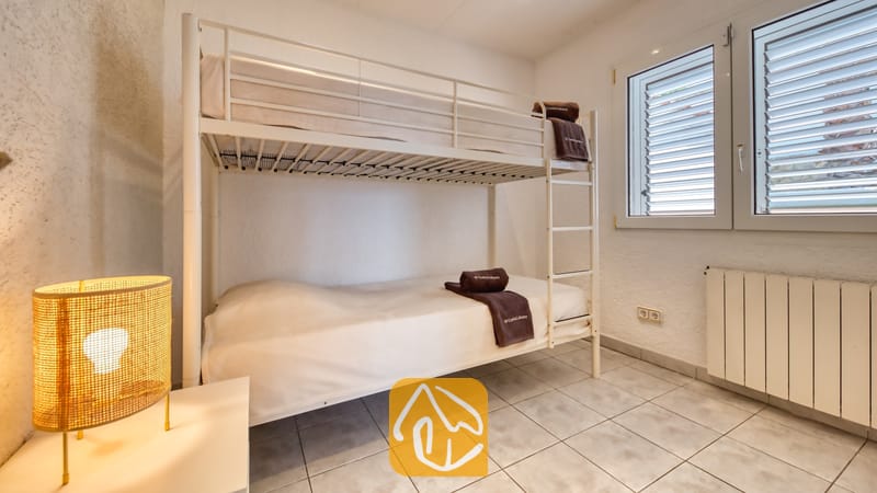 Vakantievilla's Costa Brava Spanje - Villa Maribel - Slaapkamer met een wit metalen stapelbed, een tegelvloer, een geweven lamp op een nachtkastje en twee ramen met witte luiken.