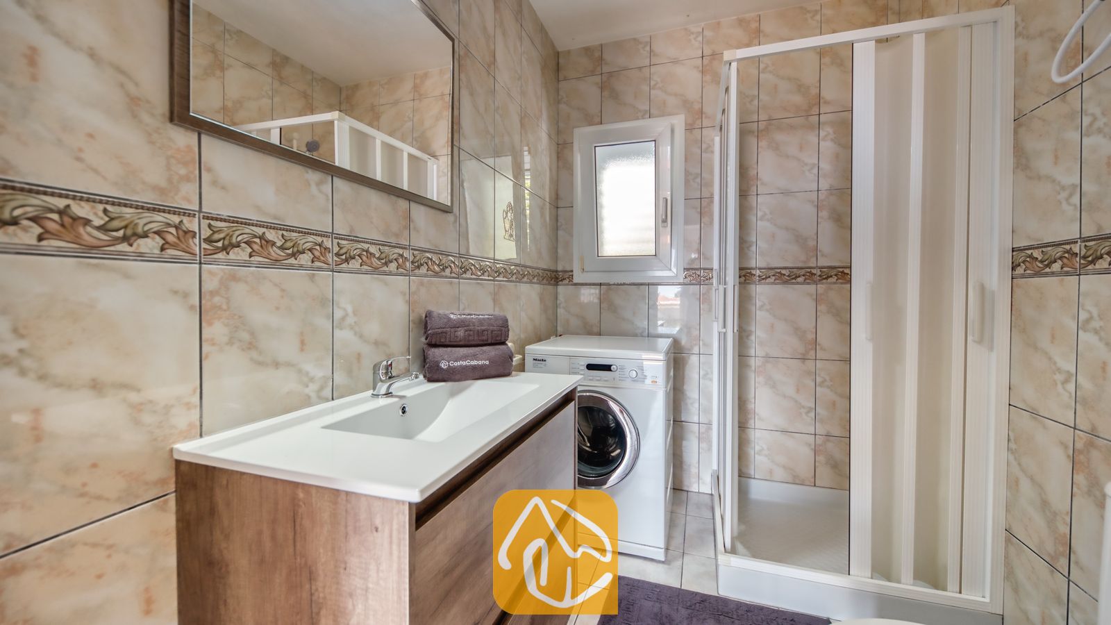 Vakantievilla's Costa Brava Spanje - Villa Maribel - Badkamer met een wit badmeubel met houten laden, een wasmachine en een glazen inloopdouche tegen een achtergrond van beige marmerlook tegels.