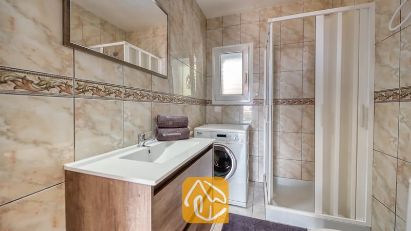 Vakantievilla's Costa Brava Spanje - Villa Maribel - Badkamer met een wit badmeubel met houten laden, een wasmachine en een glazen inloopdouche tegen een achtergrond van beige marmerlook tegels.