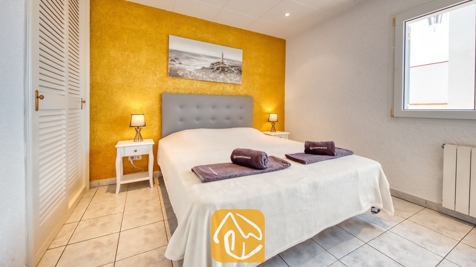Vakantievilla's Costa Brava Spanje - Villa Maribel - Slaapkamer met een tweepersoonsbed, grijs hoofdbord, opgerolde handdoeken, witte nachtkastjes met kleine lampjes en een gestructureerde gele accentmuur.