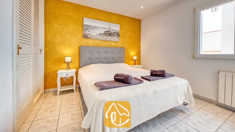 Vakantievilla's Costa Brava Spanje - Villa Maribel - Slaapkamer met een tweepersoonsbed, grijs hoofdbord, opgerolde handdoeken, witte nachtkastjes met kleine lampjes en een gestructureerde gele accentmuur.