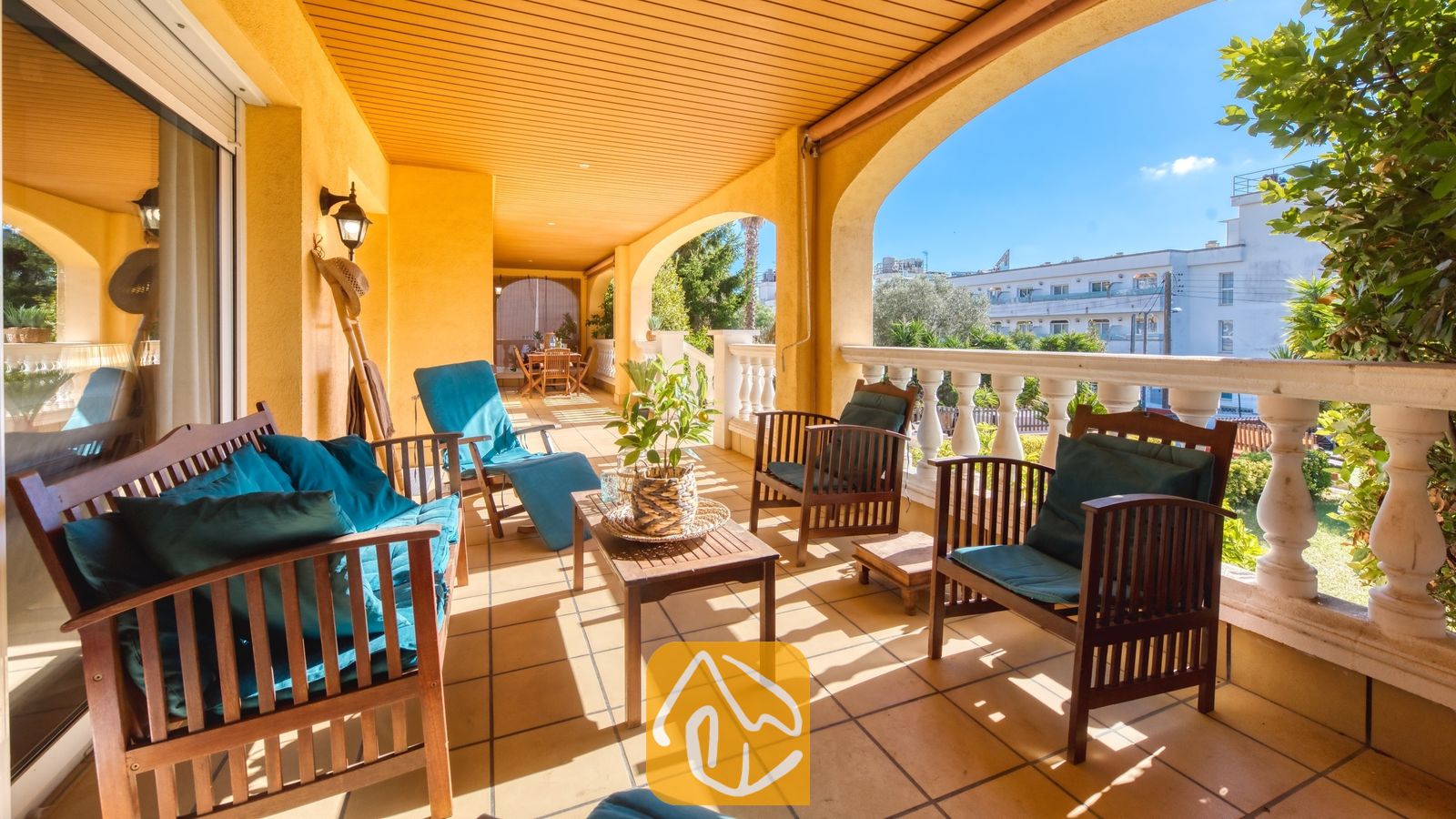 Vakantievilla's Costa Brava Spanje - Villa Summertime - Overdekt terras bij Villa Summertime met gele muren, houten zitmeubelen met the سفیدe (teal) kussens, een kleine salontafel met een potplant en een witte balustrade met uitzicht op gebouwen en bomen onder een