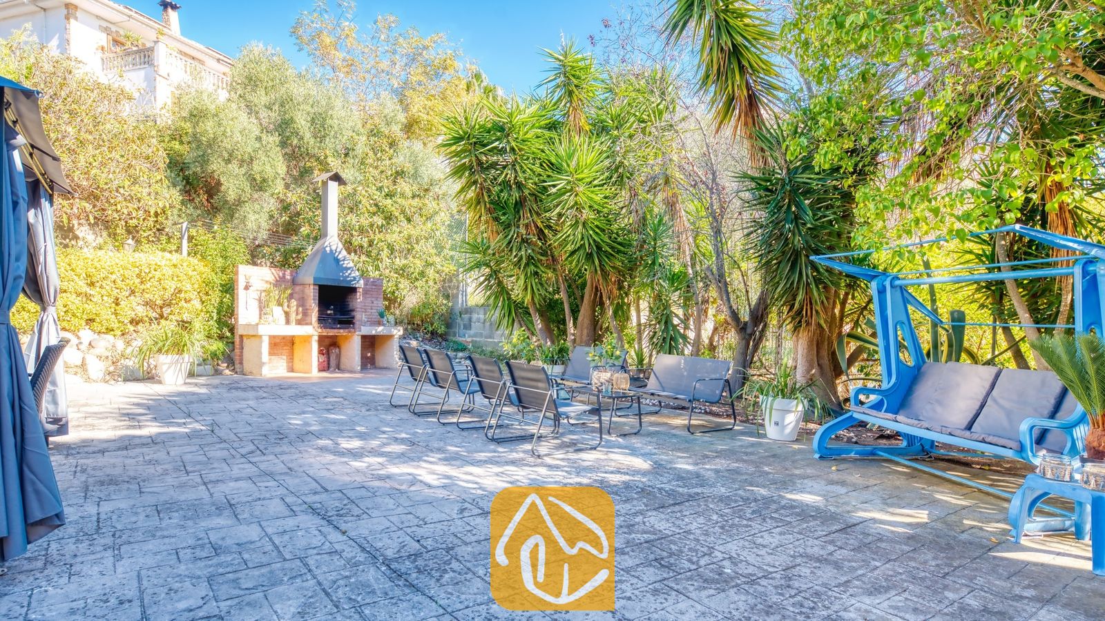 Vakantievilla's Costa Brava Spanje - Villa Summertime - Zonnig tuinterras bij Villa Summertime met een stenen barbecue, metalen tuinstoelen en een helderblauwe schommel, omringd door weelderige bomen.