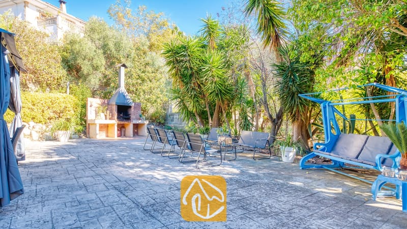 Vakantievilla's Costa Brava Spanje - Villa Summertime - Zonnig tuinterras bij Villa Summertime met een stenen barbecue, metalen tuinstoelen en een helderblauwe schommel, omringd door weelderige bomen.