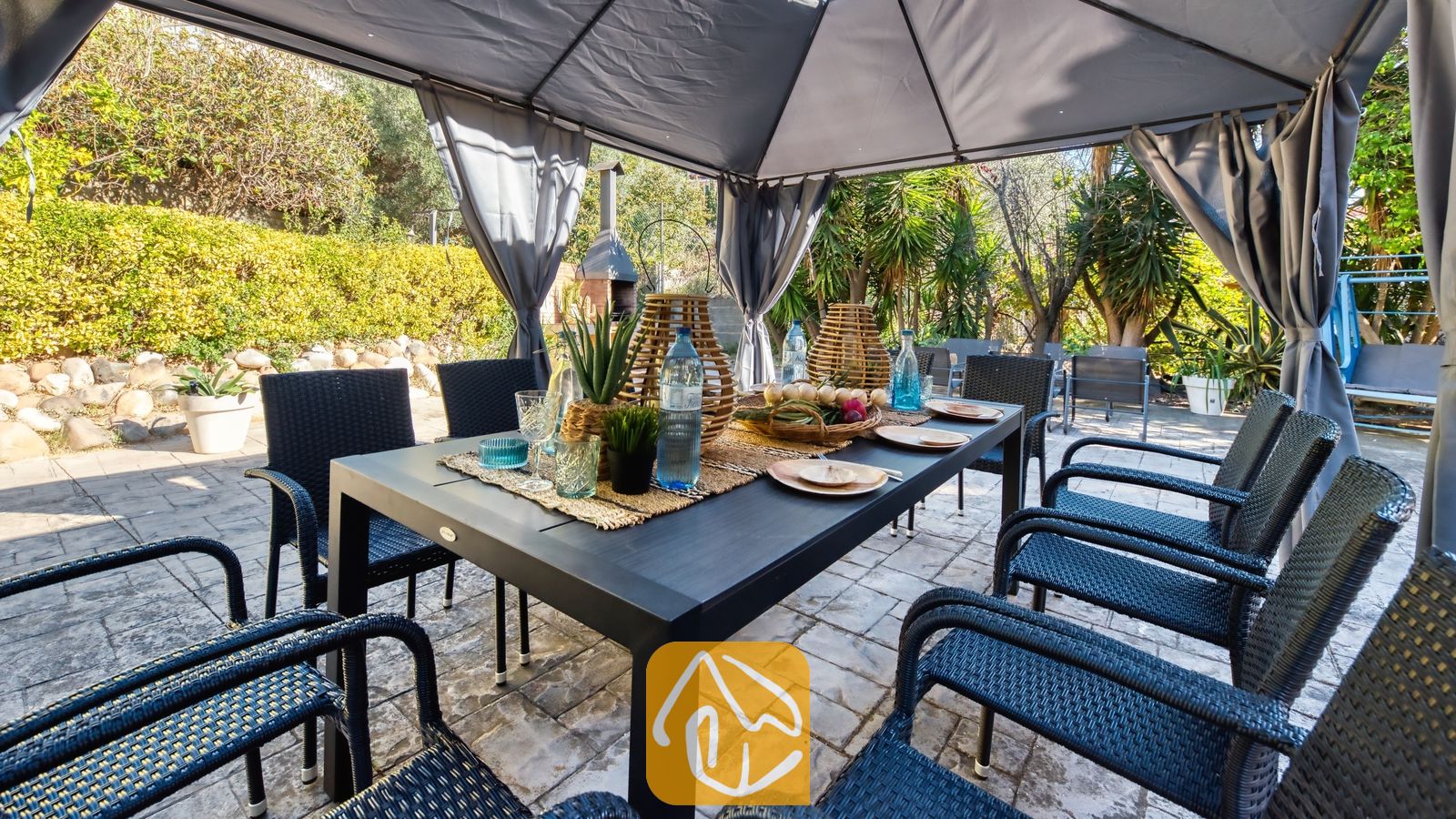 Vakantievilla's Costa Brava Spanje - Villa Summertime - Overdekte eethoek buiten bij Villa Summertime met een donkere tafel, geweven zwarte stoelen en gedekte tafels onder een grijs prieel.