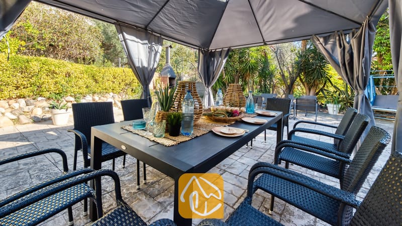 Vakantievilla's Costa Brava Spanje - Villa Summertime - Overdekte eethoek buiten bij Villa Summertime met een donkere tafel, geweven zwarte stoelen en gedekte tafels onder een grijs prieel.
