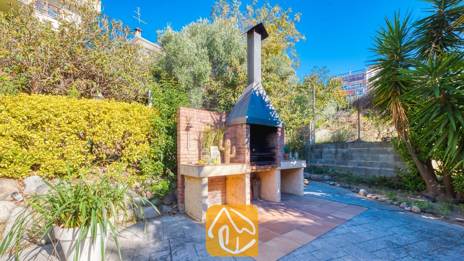 Vakantievilla's Costa Brava Spanje - Villa Summertime - Bakstenen buitenbarbecue met metalen rookkanaal in de tuin van Villa Summertime, omringd door groen loof, bomen en tegelvloeren onder een blauwe hemel.