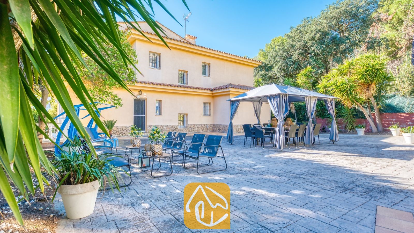 Vakantievilla's Costa Brava Spanje - Villa Summertime - Buitenterras van Villa Summertime met een stenen terras, blauwe metalen loungestoelen, potplanten en een grijze luifel over een eettafel buiten.