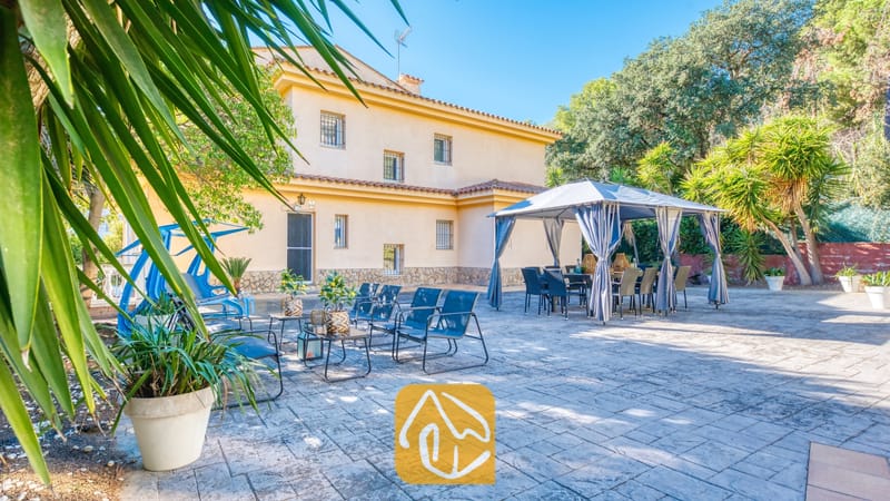Vakantievilla's Costa Brava Spanje - Villa Summertime - Buitenterras van Villa Summertime met een stenen terras, blauwe metalen loungestoelen, potplanten en een grijze luifel over een eettafel buiten.
