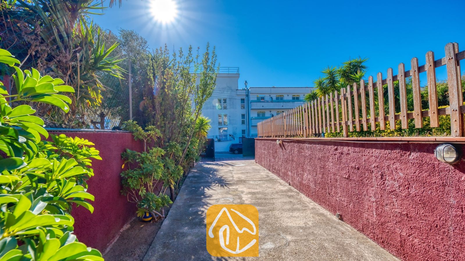 Vakantievilla's Costa Brava Spanje - Villa Summertime - Buitenpad van Villa Summertime met rode, getextureerde muren langs de rand, groene planten en een houten schutting onder een heldere, zonnige hemel.