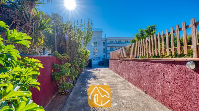 Vakantievilla's Costa Brava Spanje - Villa Summertime - Buitenpad van Villa Summertime met rode, getextureerde muren langs de rand, groene planten en een houten schutting onder een heldere, zonnige hemel.