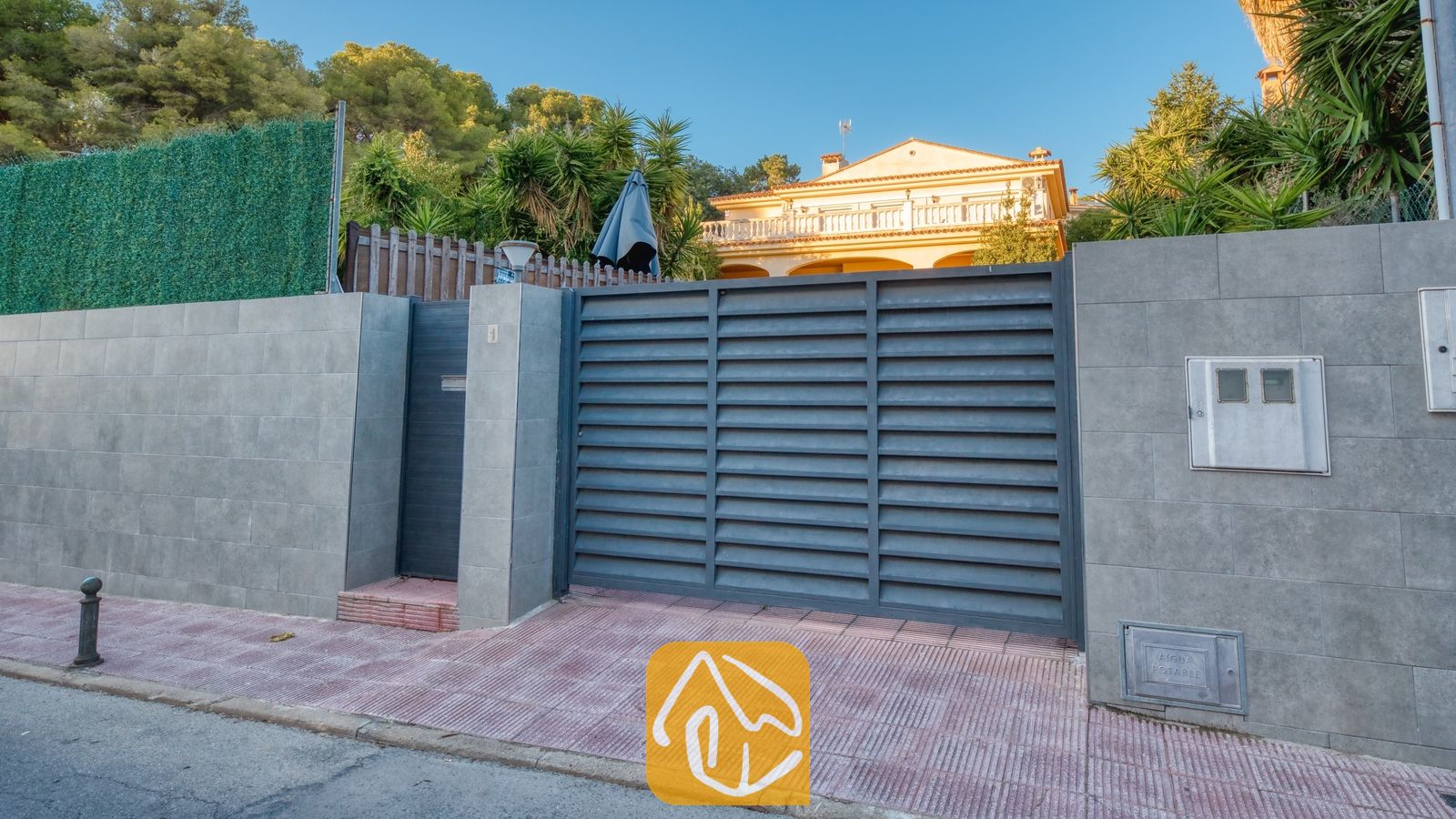 Vakantievilla's Costa Brava Spanje - Villa Summertime - Buitenkant van de entree van Villa Summertime met een grijs louvre-metalen hek en betonnen tegelmuren onder een helderblauwe lucht